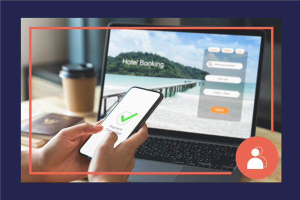 A person is holding a smart phone while using a laptop. The laptop appears to show a hotel booking website while the smart phone shows a completed payment.